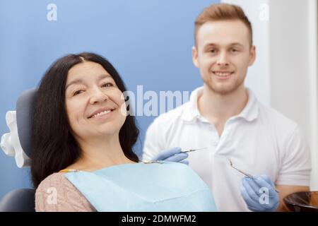 Entspannt beim Zahnarzt. Glückliche ältere Patientin sitzt in einem Zahnarztstuhl lächelnd zur Kamera ihr Zahnarzt lächelt auf dem Hintergrund hält seine Stockfoto