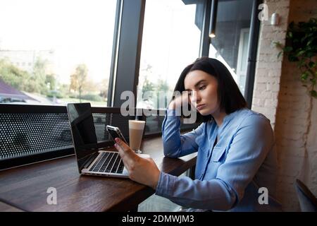 Müde Freiberuflerin, die im Café arbeitet. Konzept der Konzentrationsprobleme. Verärgert junge Frau suchen in Smartphone und wartet auf einen Anruf. Kuschelige Kaffees Stockfoto