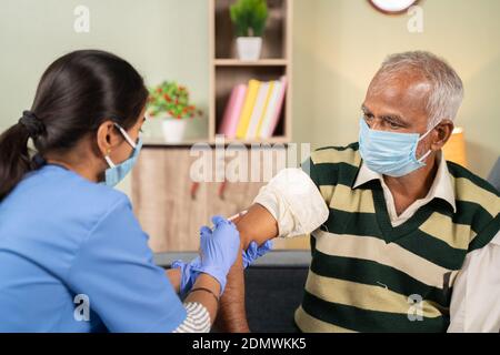 Doctor giving vaccination shot to elderly patient by syringe or injunction at home while both worj face mask - concept of home health check to seniors Stock Photo