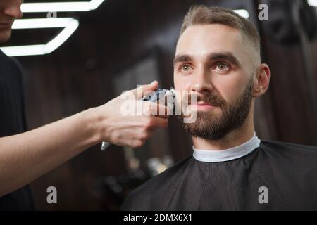 Nahaufnahme eines hübschen bärtigen Mannes, der nachdenklich wegschaut Beim Barbier mit dem elektrischen Trimmer die Seiten rasieren Stockfoto