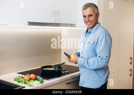 Älterer Mann, der in der Küche kocht. Fröhlicher, gut aussehender Mann mit grauen Haaren schneidet Paprika zu seinem vegetarischen Gericht hinzuzufügen. Gehacktes Gemüse in der Pfanne. Stockfoto