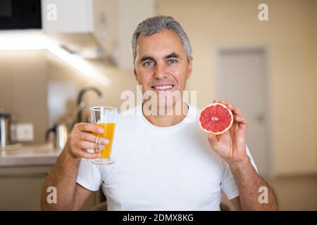 Schöner erwachsener Mann genießt sein gesundes Frühstück. Er hielt ein Glas Orangensaft und die Hälfte der Grapefruit, lächelte und schaute auf die Kamera. Vegane Küche Stockfoto