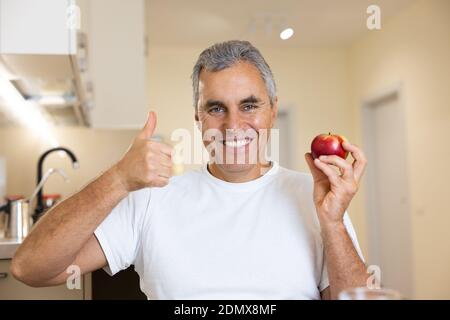 Erwachsene Mann bevorzugt gesunde Ernährung. Helles, intelligentes Interieur auf dem Hintergrund. Reifer Mann hält roten frischen Apfel, Daumen hoch und lächelt mit Zähnen. Mann sitzt im Kit Stockfoto