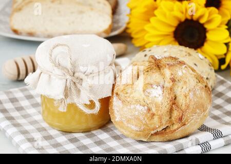 Glass jar with honey, buns and breads on breakfast table. Bouquet of sunflowers in the background. Stock Photo