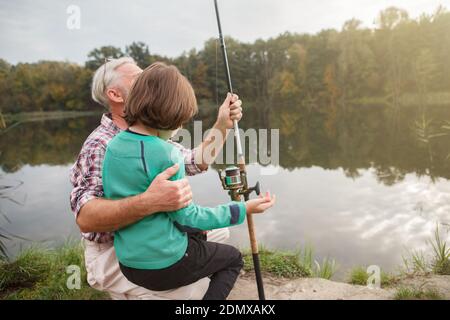 Rückansicht von einem älteren Mann umarmt seinen Enkel, während er ihn über das Angeln auf dem See, Kopierer Raum Stockfoto