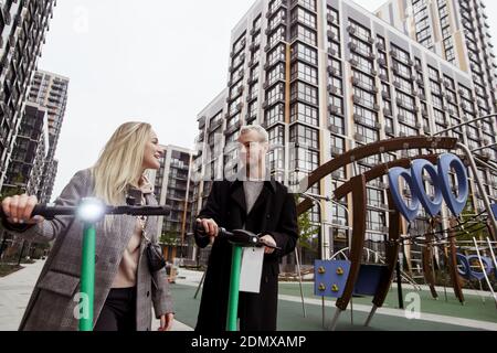 Junges Paar verbringt gute Zeit auf Elektro-Scooter. Moderne Wohnblöcke im Hintergrund. Mann und Frau mieteten E-Scooter. Schnellreisekonzept. Eco-f Stockfoto