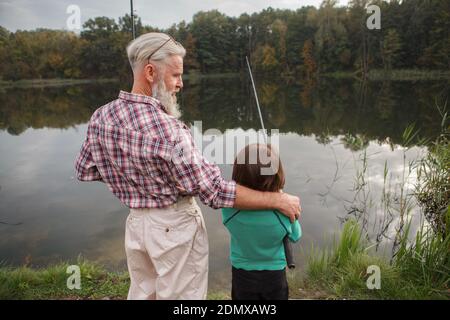 Rear view shot of a senior man hugging his grandson while the boy is fishing on the lake Stock Photo