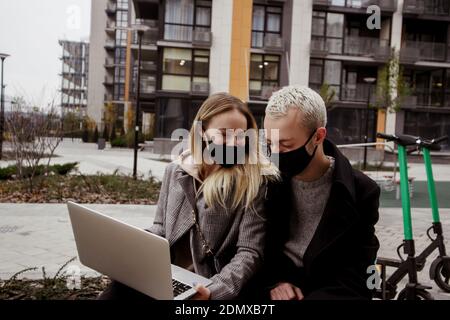Junge glückliche Paar verbringen gute Zeit auf Bank in der Nähe von Wohnblocks. Sie schauen Filme und lachen. Blonde Frau und stilvoller Mann in Gesichtsmasken halten Stockfoto