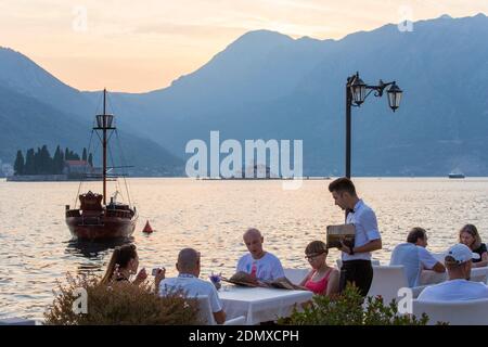 Perast, Kotor, Montenegro. Besucher, die Essen in einem Restaurant am Wasser mit Blick auf die Bucht von Kotor bestellen, Abenddämmerung. Stockfoto