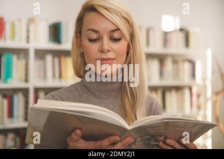 Low-Angle-Aufnahme einer schönen langhaarigen jungen Frau, die lächelt und ein Buch in der Bibliothek liest. Attraktive weibliche Prüfung Bücher, Einkaufen in den Büchern Stockfoto