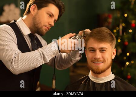 Fröhlich schöner junger bärtiger Mann lächelt freudig zur Kamera Beim Erhalten eines neuen Haarschnitts durch professionellen Barbier an der Lokaler Barbershop Copyspac Stockfoto