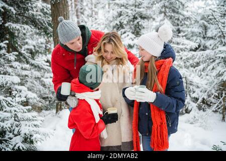 Fürsorgliche Eltern nahmen ihre Kinder mit in den Winter Park für einen Spaziergang und geben ihnen heißen Tee aus einer Thermoskanne. Gemütliches Bild im Weihnachtswald Stockfoto