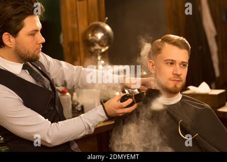 Hübscher junger Mann, der wegschaut und im Friseurstuhl sitzt Professioneller Barbier mit Talkumpuder nach der Rasur seine Kunden arbeiten Servicekontra der Mitarbeiter Stockfoto