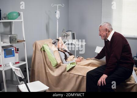 Ein Großvater jubelt seiner Enkelin im Krankenhaus zu, er erzählt ihre lustigen Geschichten, sie lacht. Stockfoto