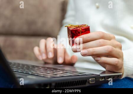 Frau in einem weißen Pullover hält einen Laptop auf dem Schoß und hält eine rote Geschenkbox in der Hand in der Nahaufnahme. Konzept von Weihnachten, Neujahr, Urlaub Stockfoto