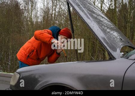 Der Mann in einer orangefarbenen Winterjacke und einem Hut spricht Am Telefon und den Motorölstand in prüfen Das Auto aus nächster Nähe Stockfoto