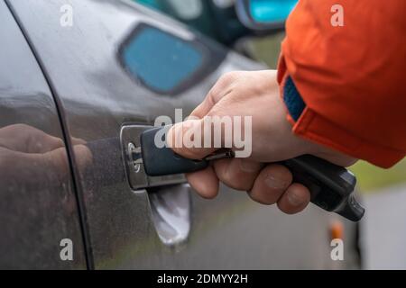 Nahaufnahme der rechten Hand eines Mannes in einer orangefarbenen Jacke, die einen Schlüssel in das Türschloss des Autos einführt. Stockfoto