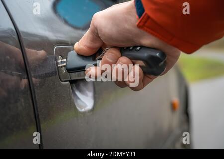 Nahaufnahme der rechten Hand eines Mannes in einer orangefarbenen Jacke, die einen Schlüssel in das Türschloss des Autos einführt. Stockfoto