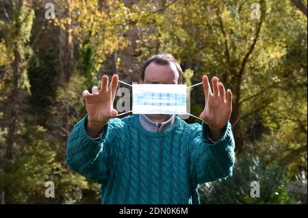 Maske von den Händen eines Mannes mit einem grünen Pullover in der Mitte der Natur gehalten. Stockfoto