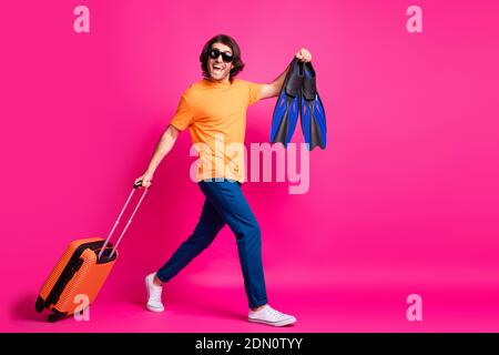 In voller Länge Profil Foto von Mann gehen halten Gepäck Flips Tragen orange T-Shirt Jeans Sonnenschutz Turnschuhe isoliert rosa Farbe Hintergrund Stockfoto