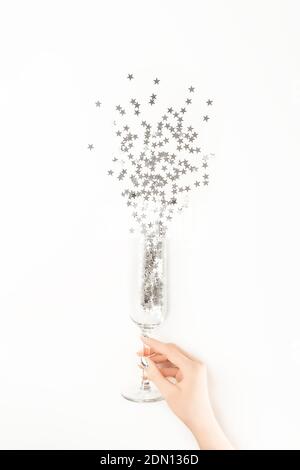 Weibliche Hand mit trendigen silbernen Nagel-Design hält Champagner-Glas mit silbernen sternförmigen Konfetti gegossen aus ihm auf weißem Hintergrund. Flacher Lay Sty Stockfoto