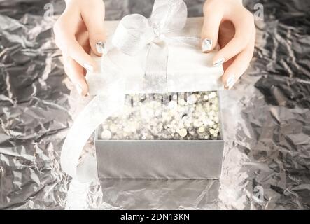 Weibliche Hände mit schönen silbernen Nagel Design Öffnung Geschenk-Box mit eleganten Bogen auf silbernen Folie Hintergrund. Weihnachtslichter leuchten in der Box Stockfoto