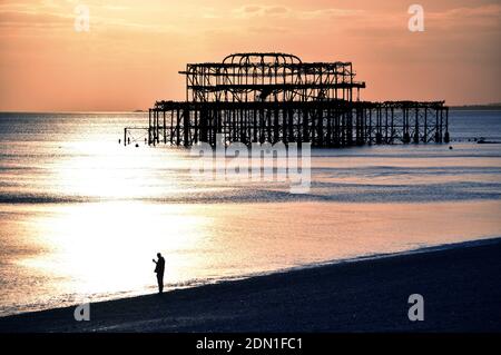 Silhouette eines einsamen Mannes mit Telefon, der bei Sonnenuntergang am Strand steht, mit Ruinen des verlassenen West Pier in Brighton, Sussex, England, Großbritannien Stockfoto