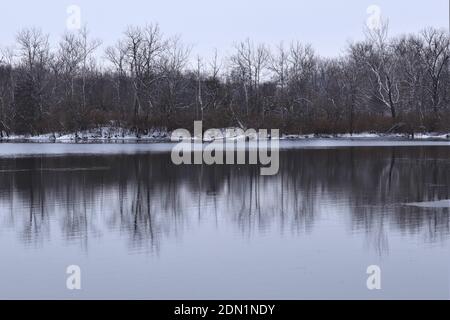 Winter landscape, lake among forest trees, trees are reflected in the water. There is snow on the ground, but the water in the lake is not frozen yet Stock Photo