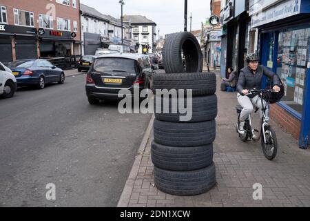 Woman cycling past a pile of tyres on the pavement outside a garage on Ladypool Road while the second national lockdown continues in Sparkhill as all non-essential shops are closed on 24th November 2020 in Birmingham, United Kingdom. The national lockdown is a huge blow to the economy and for individual businesses who were already struggling with only offering limited services. Stock Photo