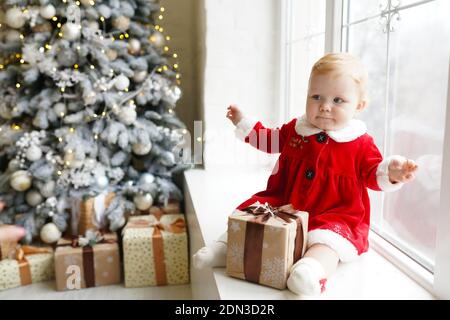 Ein kleines Rotschopf-Mädchen in einem Weihnachtskostüm in einem roten Kleid sitzt auf der Fensterbank neben weihnachtsbaum mit weihnachtsgeschenk-Box. Stockfoto