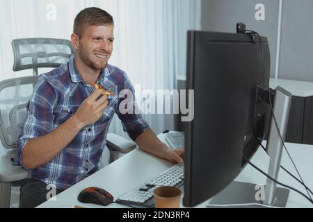 Handsome bearded IT industry worker enjoying working online, eating pizza in front of computer. Happy young man studying, eating pizza Stockfoto