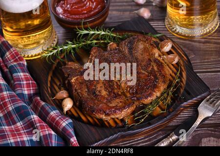 Gebratenes Schweinekotelett auf dem Knochensteak. Das Fleisch wird auf einem Holzteller mit Rosmarin und Thymian, gebratenem Knoblauch serviert. Stockfoto