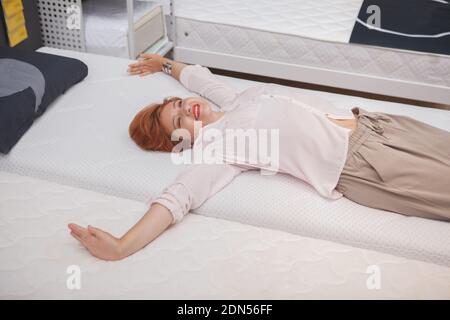 Aufgeregt junge Frau streckt ihren Rücken liegend auf orthopädische Matratze Im Möbelgeschäft Stockfoto