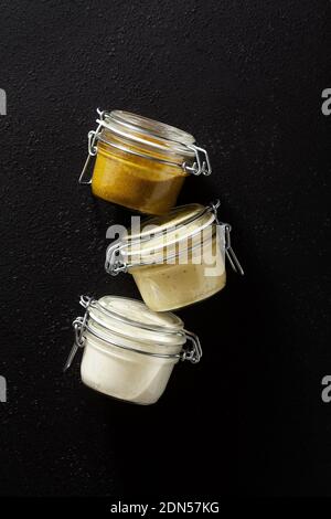 different types of sauces in glass jars. with pistachios, cashews and vegan mayo on black background Stockfoto