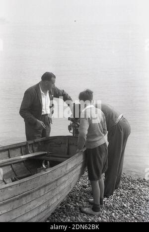 1950er, historisch, zwei Männer und ein Jugendlicher stehen an einem hölzernen Ruderboot am Steinstrand, am Meeresrand bei Walmer, Deal, Kent, England, Großbritannien, Mit den Erwachsenen, um den Außenbordmotor zu überprüfen. Vielleicht brauchen sie es, denn die Wasserfläche vor ihnen ist der Ärmelkanal. Stockfoto