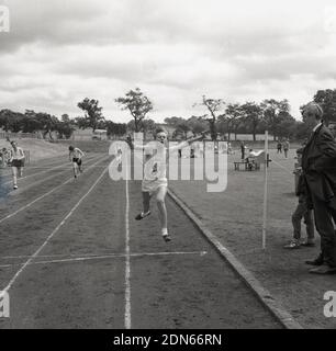 1960er, historische, junge Männer, die an einem Laufrennen auf einer Aschebahn teilnehmen, an einem County-Sporttag in Fife, Schottland, mit einem jungen Mann, der sich die Arme vor dem Band ausstreckt. Stockfoto