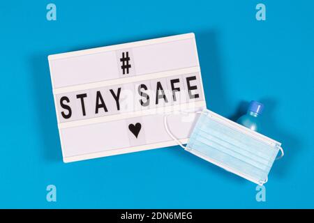 Weißer Leuchtkasten mit Meldung Stay Safe, medizinische Schutzmaske und Handgel auf hellblauem Hintergrund. Draufsicht Stockfoto