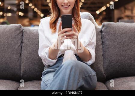 Ausgeschnittene Aufnahme einer lächelnden Frau, die Smartphone benutzt, während sie sich auf einem Sofa ausruhte Stockfoto