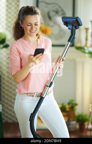 Happy Modern 40 Jahre alte Frau in Bluse und weißen Hosen mit Staubsauger mit Home Cleaning App auf dem Smartphone im modernen Zuhause an sonnigen Tagen. Stockfoto