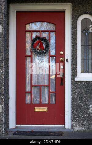 Traditioneller grüner Weihnachtskranz, der an der roten Haustür hängt Eines Hauses Stockfoto