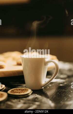 Vertikales Foto einer weißen Tasse mit etwas Dampf Ein schwarzer Tisch in der Nähe einer Holzplatte und einige getrocknet Limetten Stockfoto