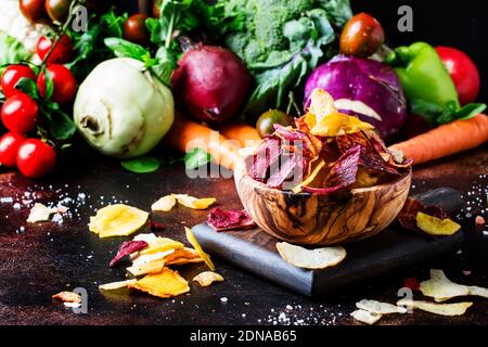 Vegane Snacks, bunte Gemüsechips in Holzschüssel und Set aus frischem Bauerngemüse, rustikales Stillleben, selektiver Fokus Stockfoto