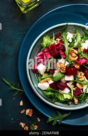 Rüben- oder Rote Bete-Salat mit frischem Rucola, Radicchio, Weichkäse und Walnüssen auf Teller mit Gabel, Dressing und Gewürzen auf blauem Küchentischhintergrund Stockfoto