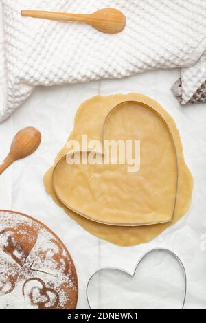 Eine herzförmige Schneideform auf gerolltem Teig neben einem Handtuch und Holzlöffeln. Flach liegend. Vertikal Stockfoto