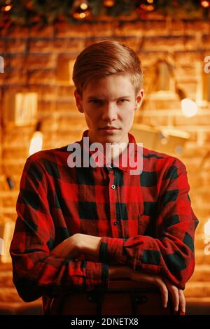 Serious Kerl auf Hochstuhl in Weihnachtsdekorationen. Ein hübscher Mann im karierten Hemd blickt unter seiner Stirn auf die Kamera. Die Atmosphäre des neuen Jahres. Backsteinmauer mit festlicher Einrichtung im Hintergrund. Stockfoto