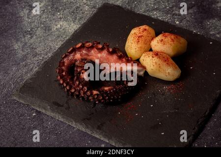 Gegrillte Tintenfisch-Tentakeln mit Kartoffeln gewürzt mit spanischem Paprika, Olivenöl und Meersalz auf einer schwarzen Platte und schickem Hintergrund. Gourmet Küche CO Stockfoto