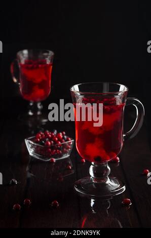 Roter Früchtetee mit Preiselbeere, Heidelbeere in zwei transparenten Gläsern auf schwarzem Holztisch im dunklen Innenraum, senkrecht. Stockfoto