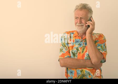 Studio shot der älteren bärtigen touristische Mann betonte, beim Denken und Sprechen auf Handy vor weißem Hintergrund suchen Stockfoto