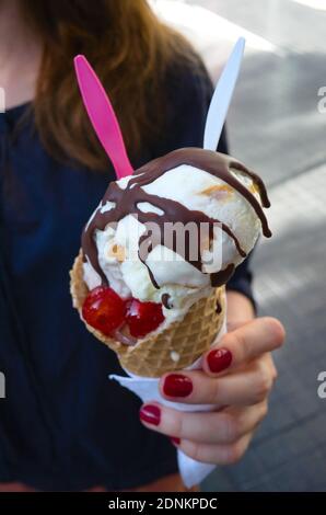 Eis in weiblichen Händen Nahaufnahme. Hand mit roten Nägeln hängen Eis mit Schokolade, Kirschen und zwei Löffel im Freien auf der Straße. Stockfoto