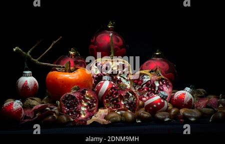 Granatapfel offen mit Samen und eine Kaki mit einem geschmückt Rote Weihnachtskugeln auf Kastanien vor schwarzem Hintergrund Stockfoto